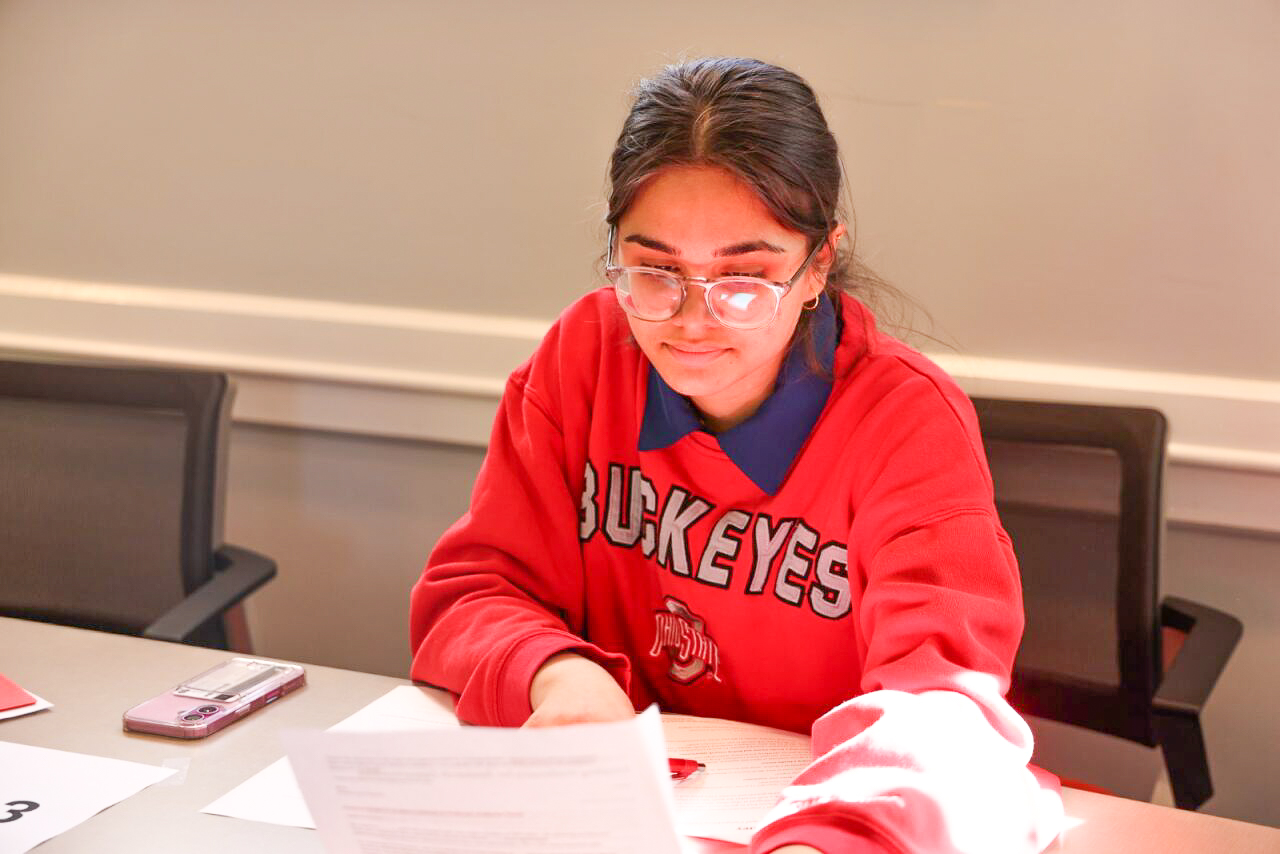 A person sitting at a table, looking at papers. They are wearing glasses and a red sweatshirt with "Buckeyes" on it.