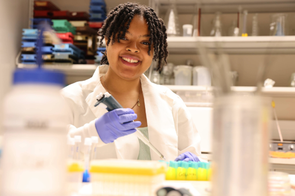A person wearing a white lab coat and gloves is using a pipette in a laboratory. Test tubes and various lab equipment are visible 