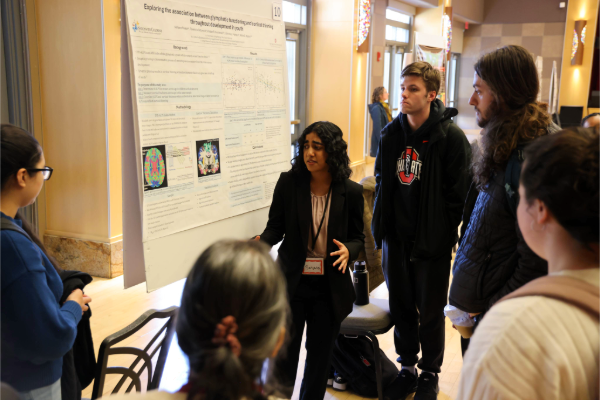 A person stands in front of a research poster, engaged in explaining the content to a small group of attentive individuals.