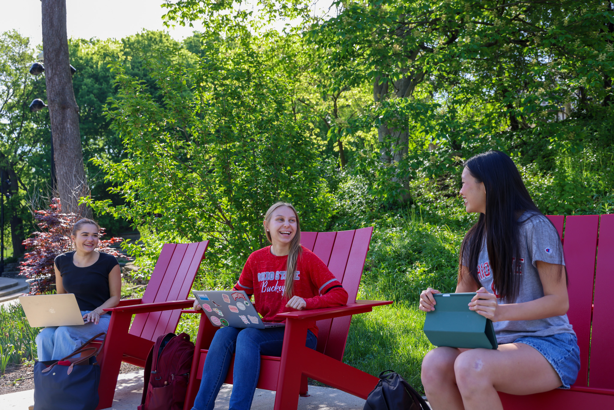 Three people sitting outdoors in red chairs are laughing and engaged in conversation.