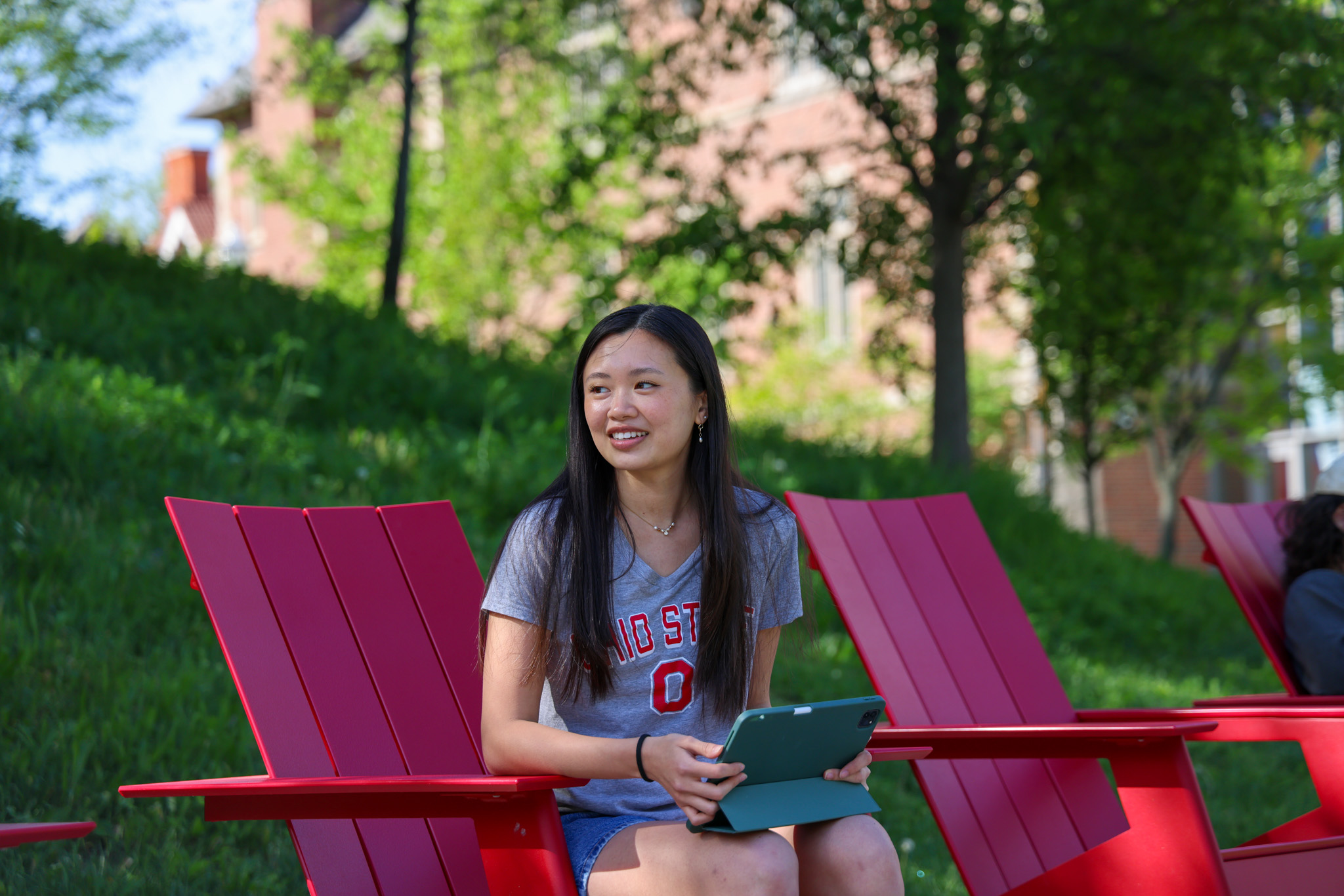 A person is sitting on a red outdoor chair, holding a tablet and smiling wearing an Ohio State t-shirt.