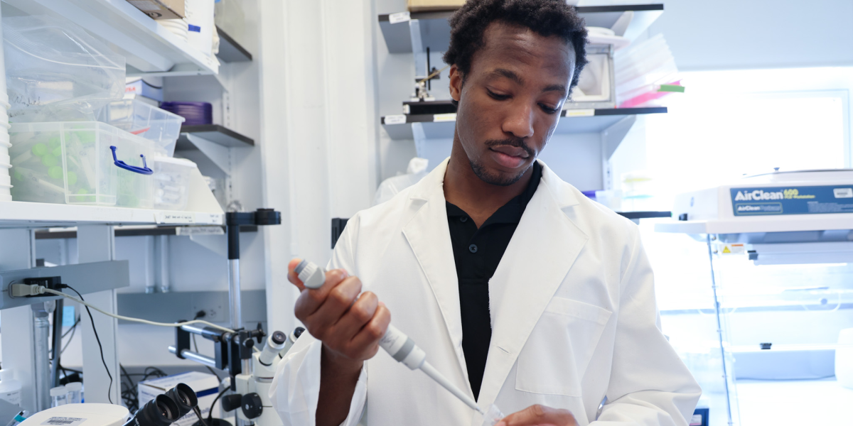A person in a lab coat holds a pipette in a laboratory setting, standing near microscopes and equipment, including an AirClean Systems apparatus.