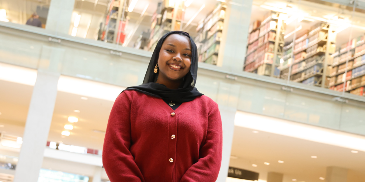 Daranii Asoba in a red cardigan stands smiling in a well-lit library with multiple shelves filled with books in the background.
