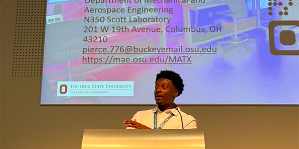 Malcolm Pierce stands at a podium giving a presentation. Behind is a slide displaying information