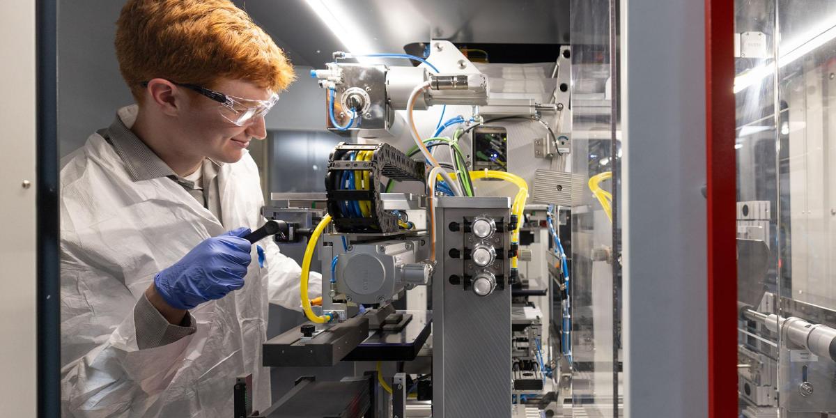 Individual smiles while operating battery wearing a lab coat, goggles, and gloves in lab.