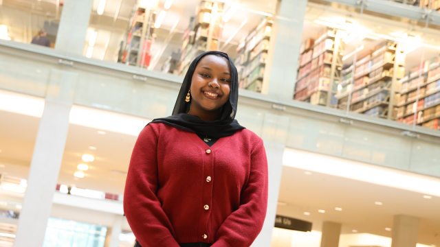 Daranii Asoba in a red cardigan stands smiling in a well-lit library with multiple shelves filled with books in the background.
