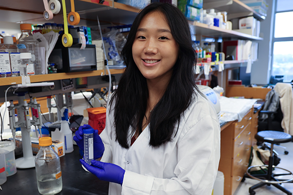 A person wearing a lab coat and gloves holds a test tube in a laboratory.