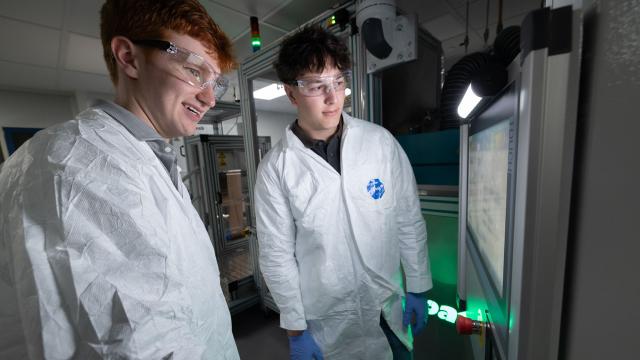 Two individuals stand in lab coats and goggles looking at screen