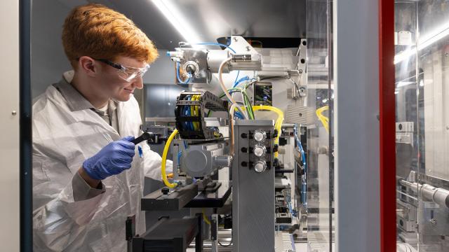 Individual smiles while operating battery wearing a lab coat, goggles, and gloves in lab.