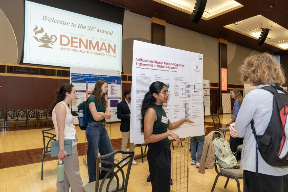 A group of individuals are engaged in discussions around research posters with Denman logo in background