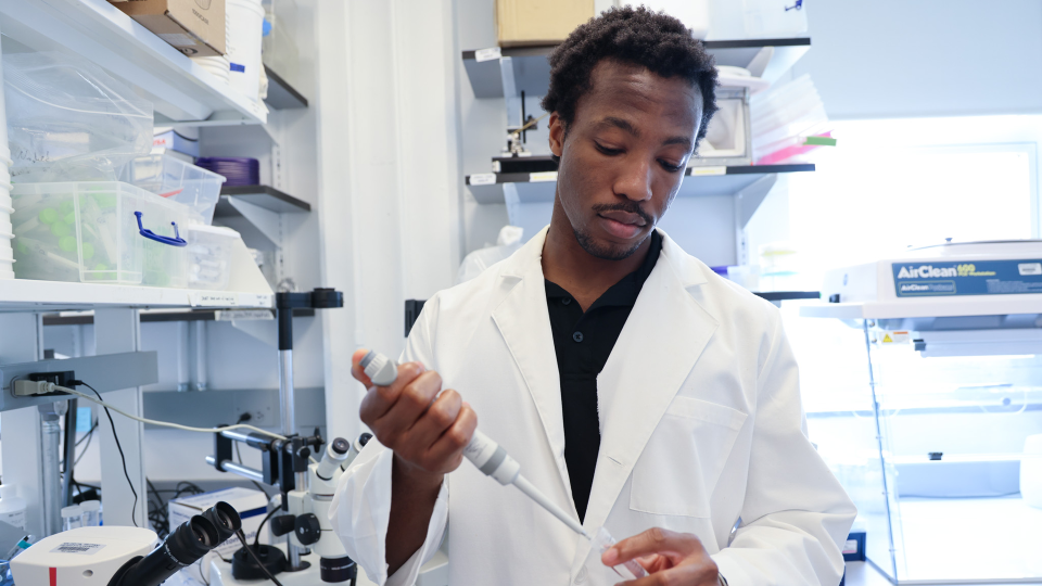 A person in a lab coat holds a pipette in a laboratory setting, standing near microscopes and equipment, including an AirClean Systems apparatus.