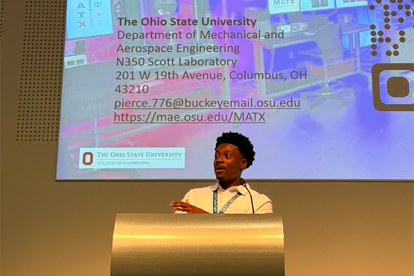 Malcolm Pierce stands at a podium giving a presentation. Behind is a slide displaying information