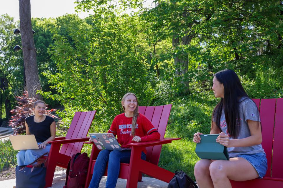 Three people sitting outdoors in red chairs are laughing and engaged in conversation.