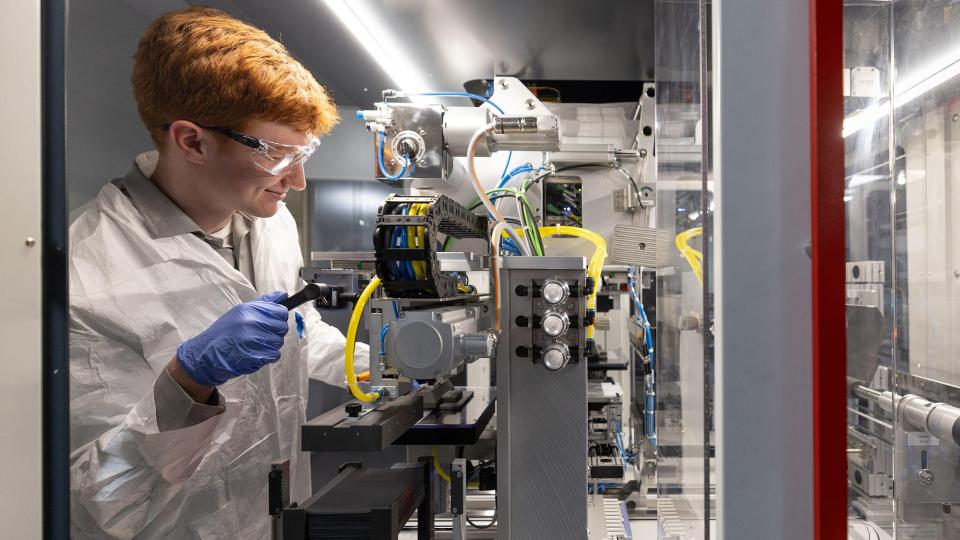 Individual smiles while operating battery wearing a lab coat, goggles, and gloves in lab.