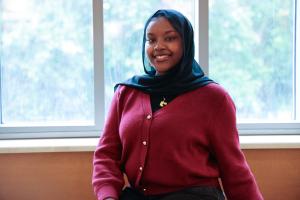 Daranii Asoba wearing a red cardigan with gold buttons and a dark headscarf, sitting in front of a large window with a blurred background of greenery.