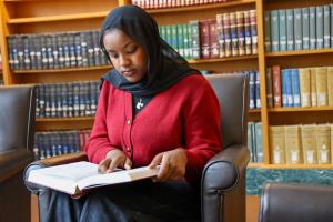 A person is sitting in a library reading a book, surrounded by shelves filled with books.