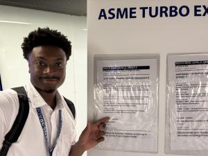 Malcolm Pierce stands next to a board labeled "ASME TURBO EXPO" and pointing at printed notices related to conference sessions.