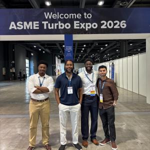 Four people are standing together smiling in front of a sign that reads "Welcome to ASME Turbo Expo 2026" in a large convention center.