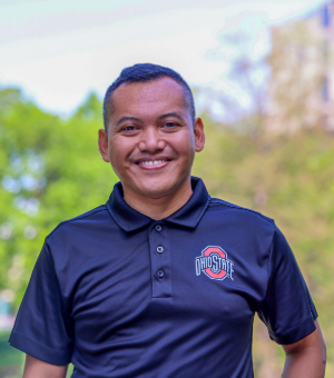 A person wearing a black polo shirt with the Ohio State University logo stands outdoors, smiling. The background is a blurred mix of greenery and buildings.
